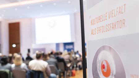 Am rechten Bildrand ist der Ausschnitt eines Banners mit dem Logo "Interkultureller Wirtschaftspreis" zu sehen. Links ist verschwommen ein Publikum im Hintergrund zu erahnen.