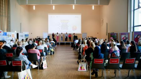 Blick auf die Bühne eines Saals, davor sitzen zahlreiche Menschen in aufgereihten Stuhlreihen. An der Bühnenrückwand ist eine Projektion eines Beamers zu sehen.